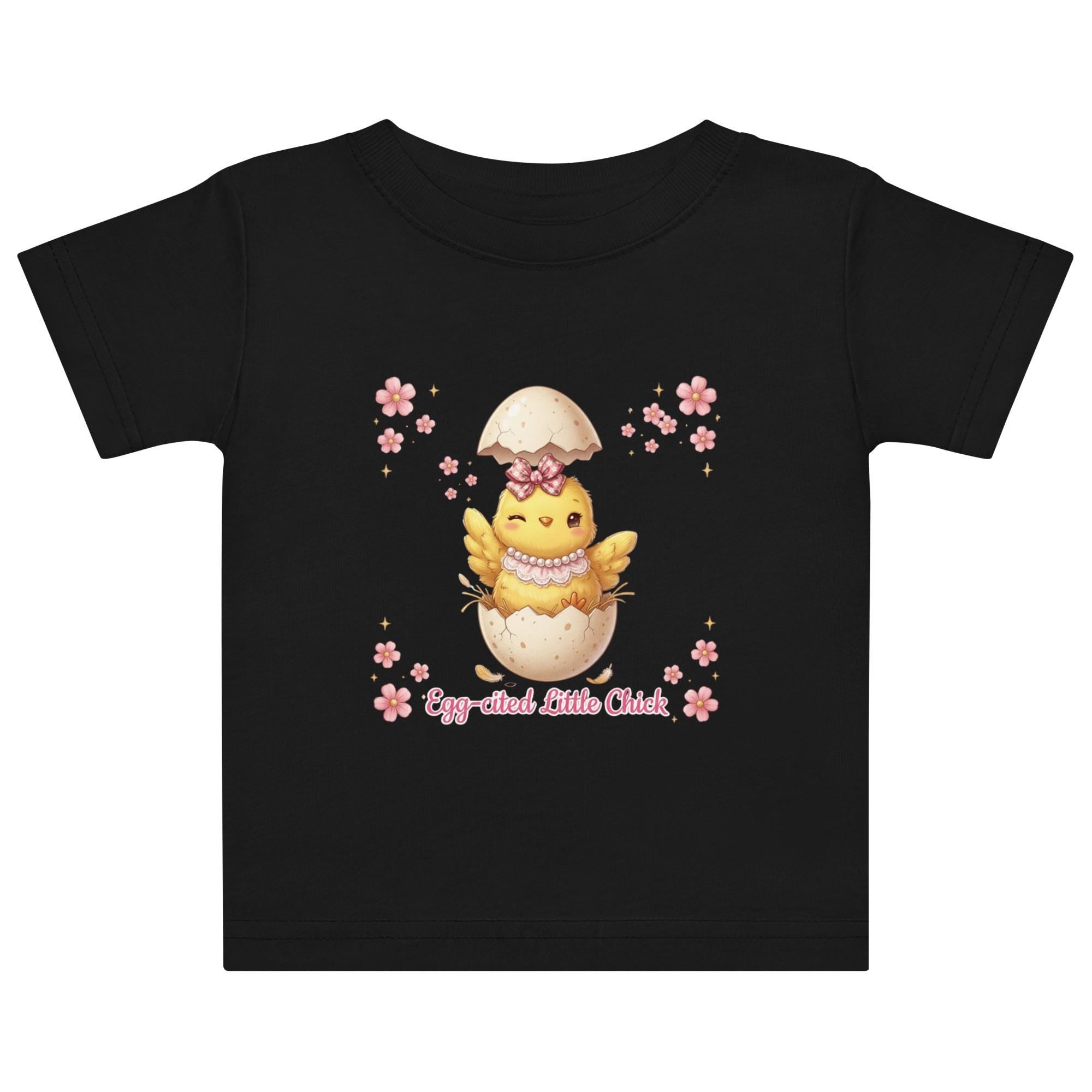 May include: White short-sleeved t-shirt featuring a cartoon chick hatching from an egg, with the text "Egg-cited Little Chick" in pink. The chick is yellow with a pink bow and necklace. The child is wearing a pink bow.