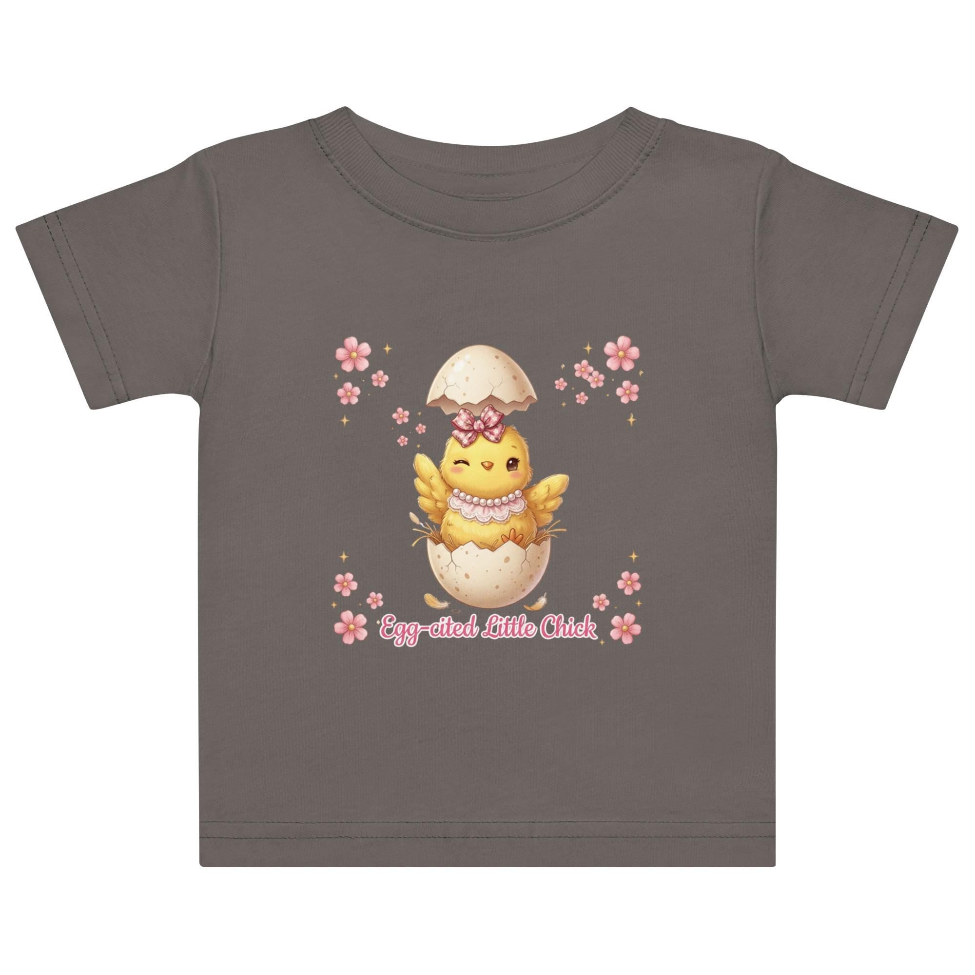 May include: White short-sleeved t-shirt featuring a cartoon chick hatching from an egg, with the text "Egg-cited Little Chick" in pink. The chick is yellow with a pink bow and necklace. The child is wearing a pink bow.