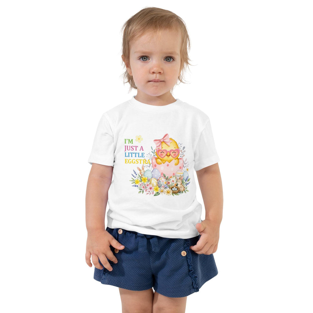 May include: A young child wearing a white t-shirt with the text "I'M JUST A LITTLE EGGSTRA" and a graphic of a chick hatching from an egg. The child is also wearing a pink tutu, pink sneakers, and white socks. The child is raising their right hand.