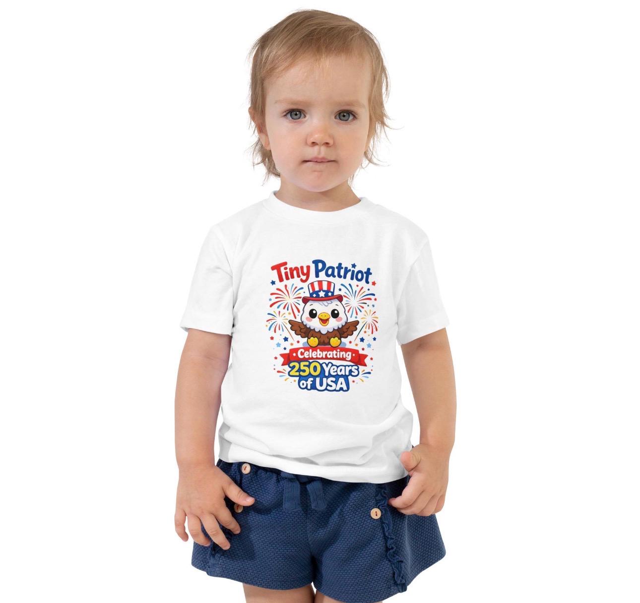 May include: A white toddler t-shirt with a colorful graphic that reads "Tiny Patriot" above an illustration of an owl wearing a patriotic hat. The graphic also includes the text "Celebrating 250 Years of USA" with fireworks.