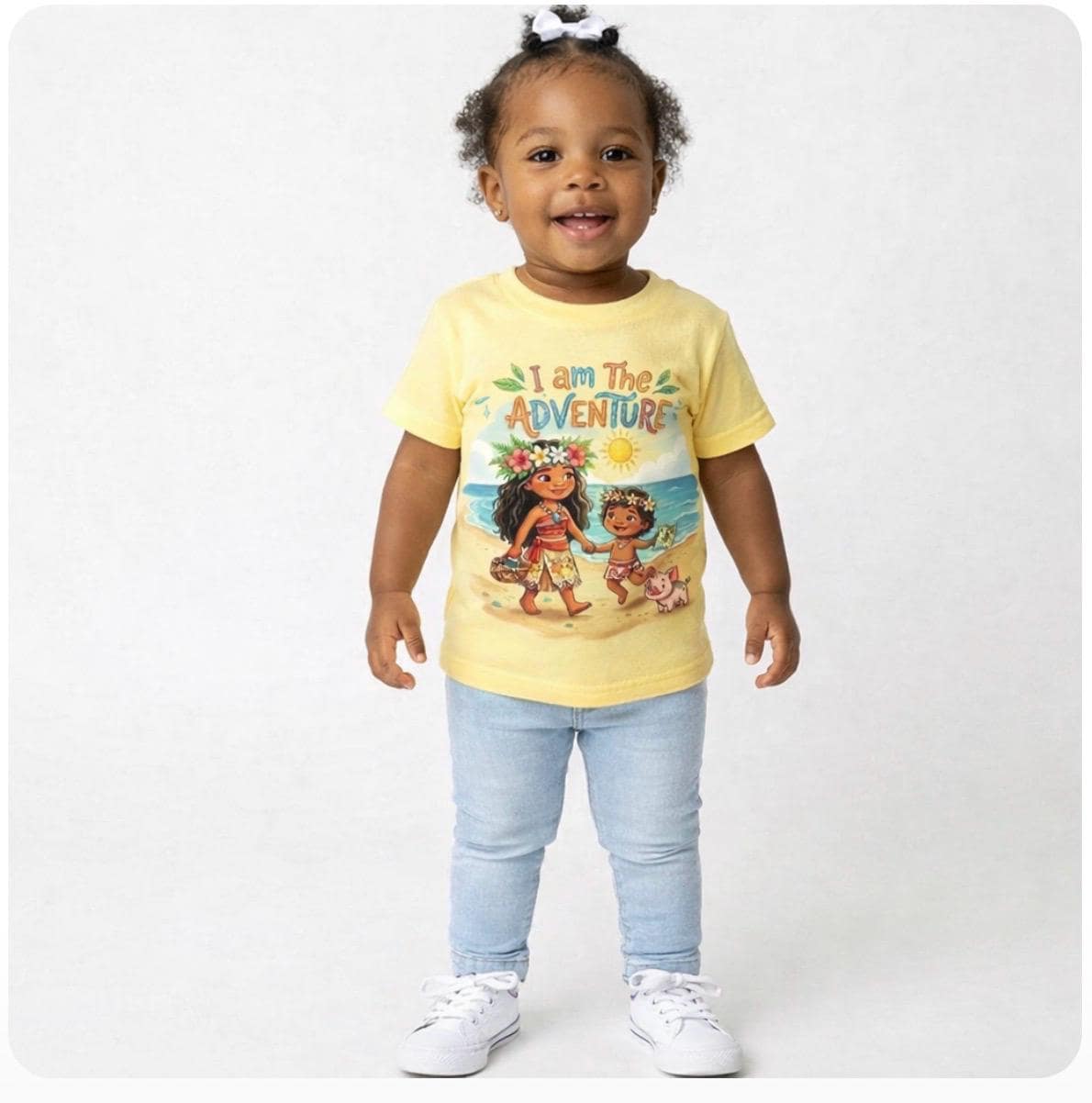 May include: A toddler wearing a yellow t-shirt with the text "I am The ADVENTURE" and a graphic of two cartoon characters on a beach. The toddler is also wearing light blue jeans and white sneakers.